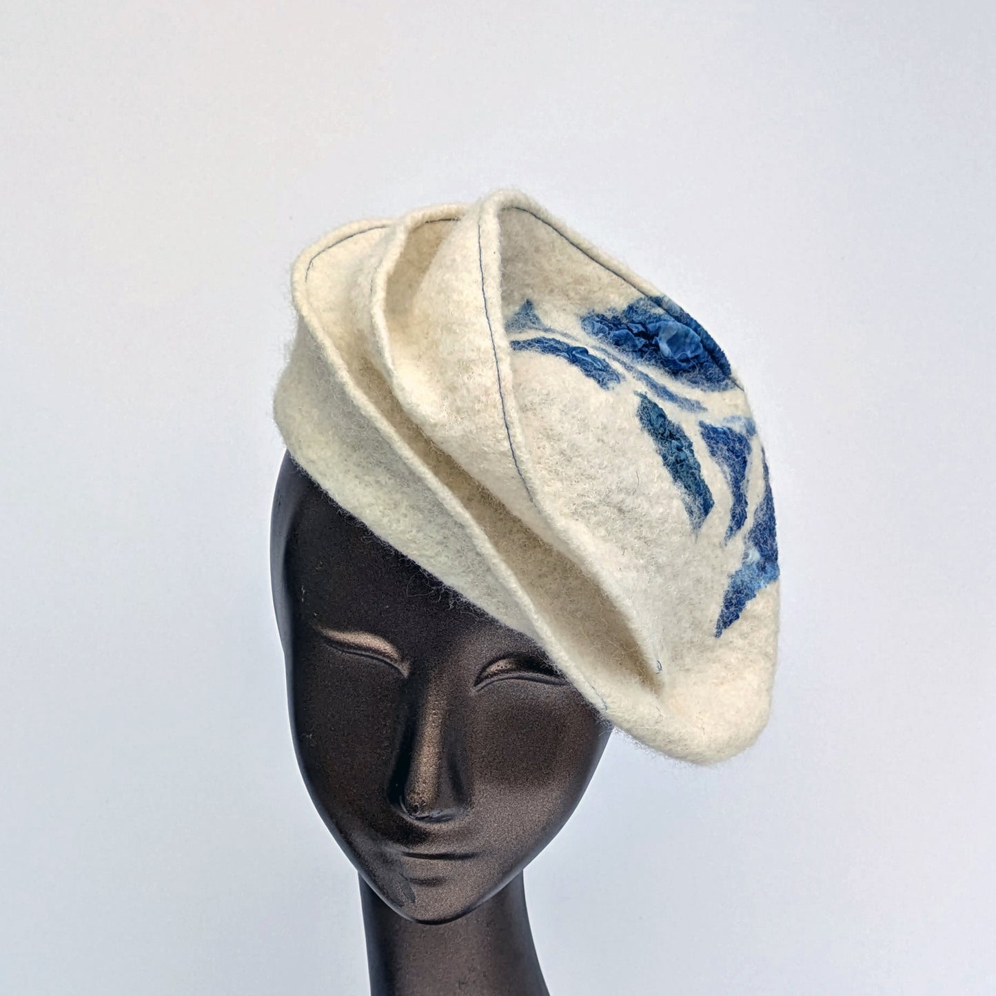 Small cream-coloured hat with blue stitchig and blue geometric patterning.