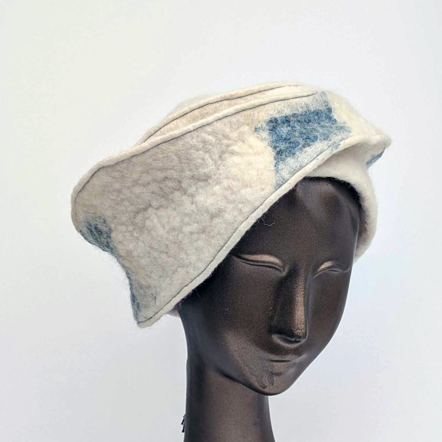 Cream and blue patterned, asymmetrical hat on a mannequin head against a white background