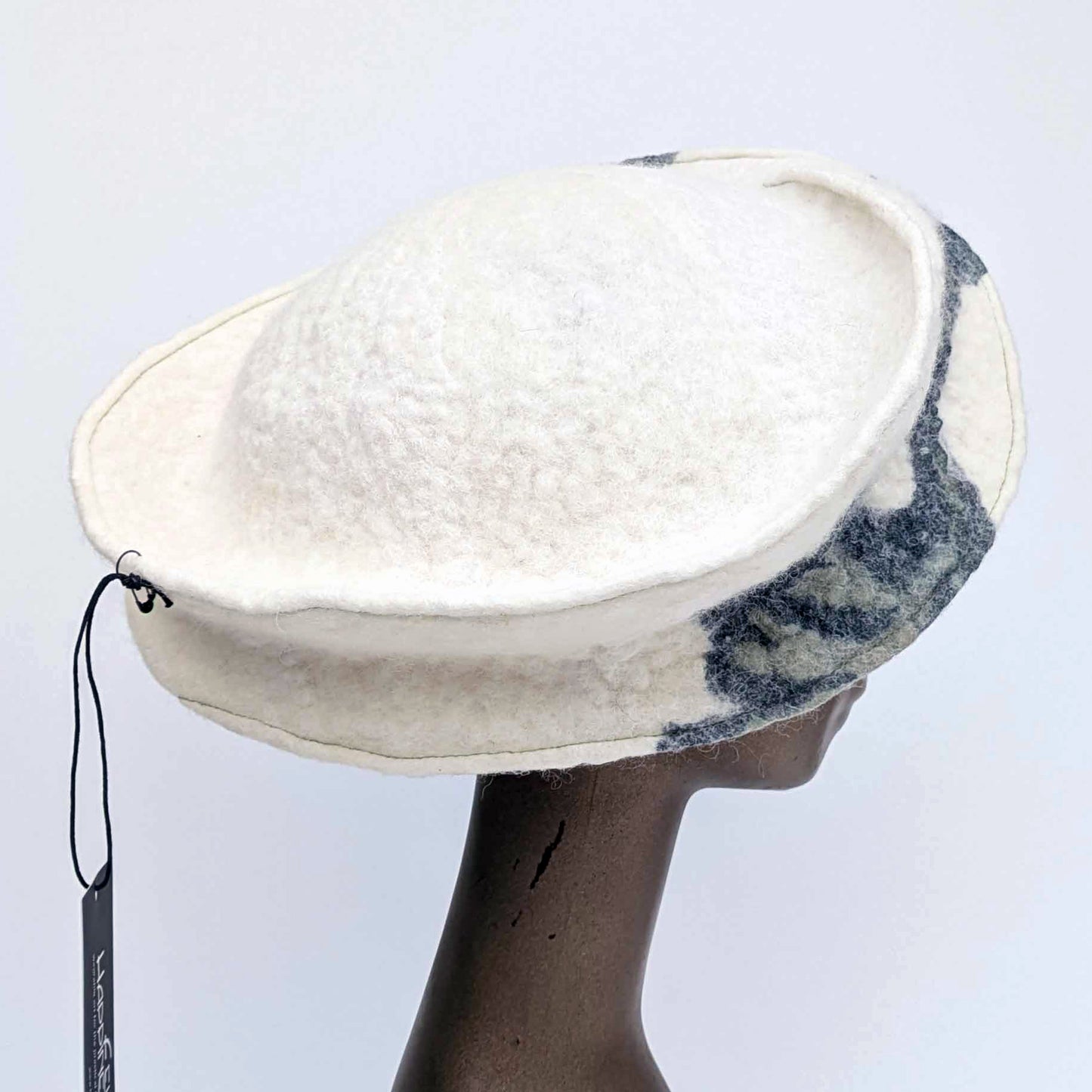 Cream coloured, asymmetrical hat with a broad band, with a small amount of dark blue decorations on the side.