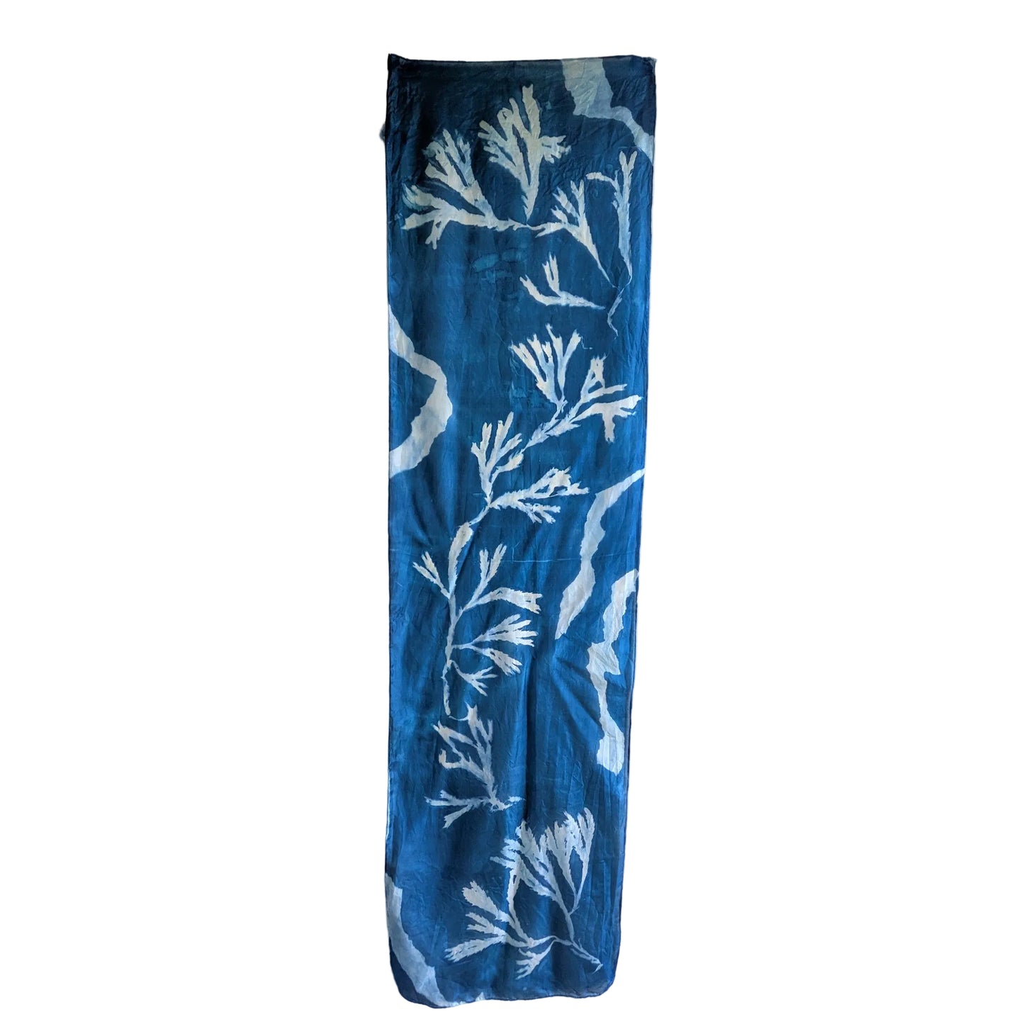 A long, narrow blue scarf hanging vertically against a plain off-white wall, featuring a white sunprint pattern made from seaweed and torn paper.