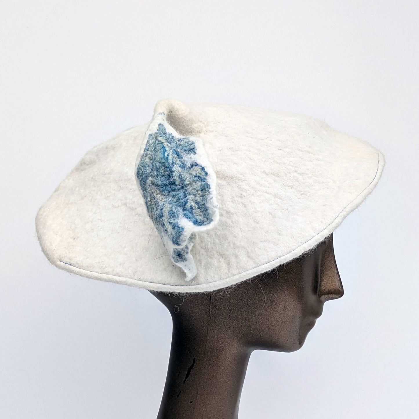 Mostly white beret with a touch of blue on the leafy tassel - worn by mannequin.