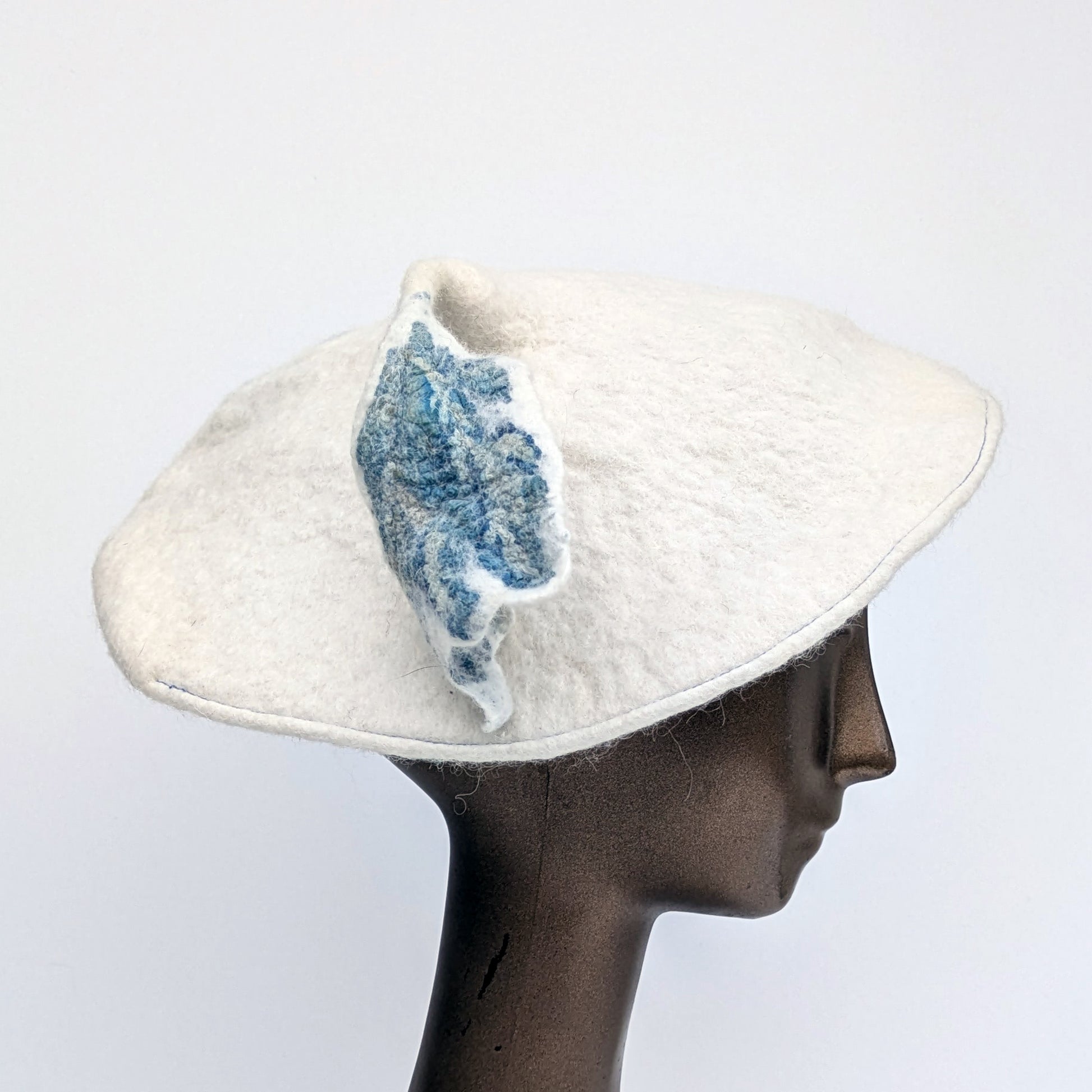 Mostly white beret with a touch of blue on the leafy tassel - worn by mannequin.