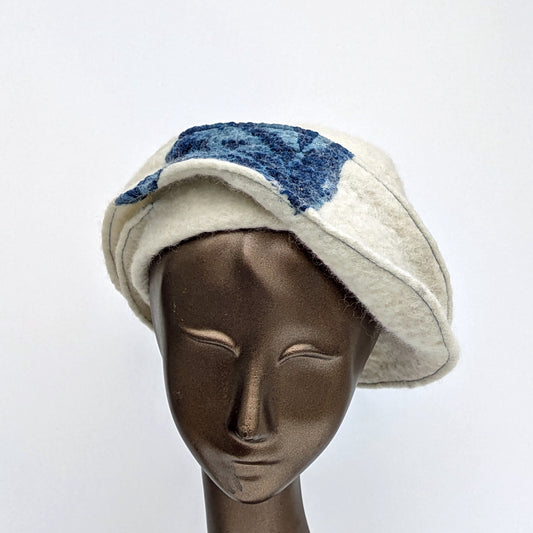 Creamy white felted hat with dark blue silk fabric. Has a small bill like a cap, which is worn to one side, asymetrically.
