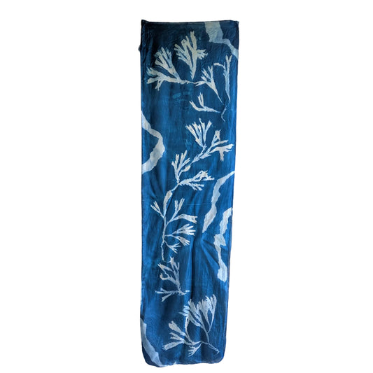 A long, narrow blue scarf hanging vertically against a plain off-white wall, featuring a white sunprint pattern made from seaweed and torn paper.