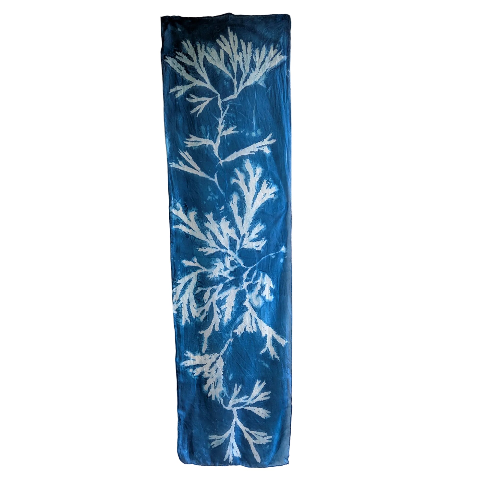 A vertical rectangular silk scarf, featuring a white seaweed sunprint pattern on a deep blue background, hanging against a plain off-white wall.